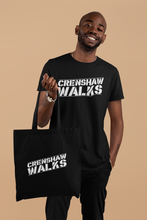 Load image into Gallery viewer, African man wearing black short sleeve tee with Crenshaw Walks across the front. The man is holding a matching black tote bag. The Crenshaw Walks design is done in a stencil style and slightly slanted up.